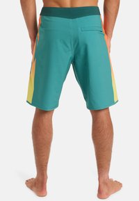 Bañadores turquesas con patrones laterales en naranja y amarillo, cinturilla elástica y un bolsillo trasero. Fabricados con material de secado rápido.