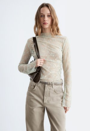 Junge Frau trägt ein durchsichtiges, langärmliges Oberteil mit Blumenmuster, beige hochtaillierte Jeans, hält eine kleine dunkelbraune Umhängetasche vor hellem Hintergrund.