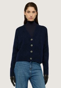 Námornícky modrý zkrácený cardigan s texturovaným vzorem, pět knoflíků vpředu a široké rukávy, ve spojení s modrými džíny a černými rukavicemi.