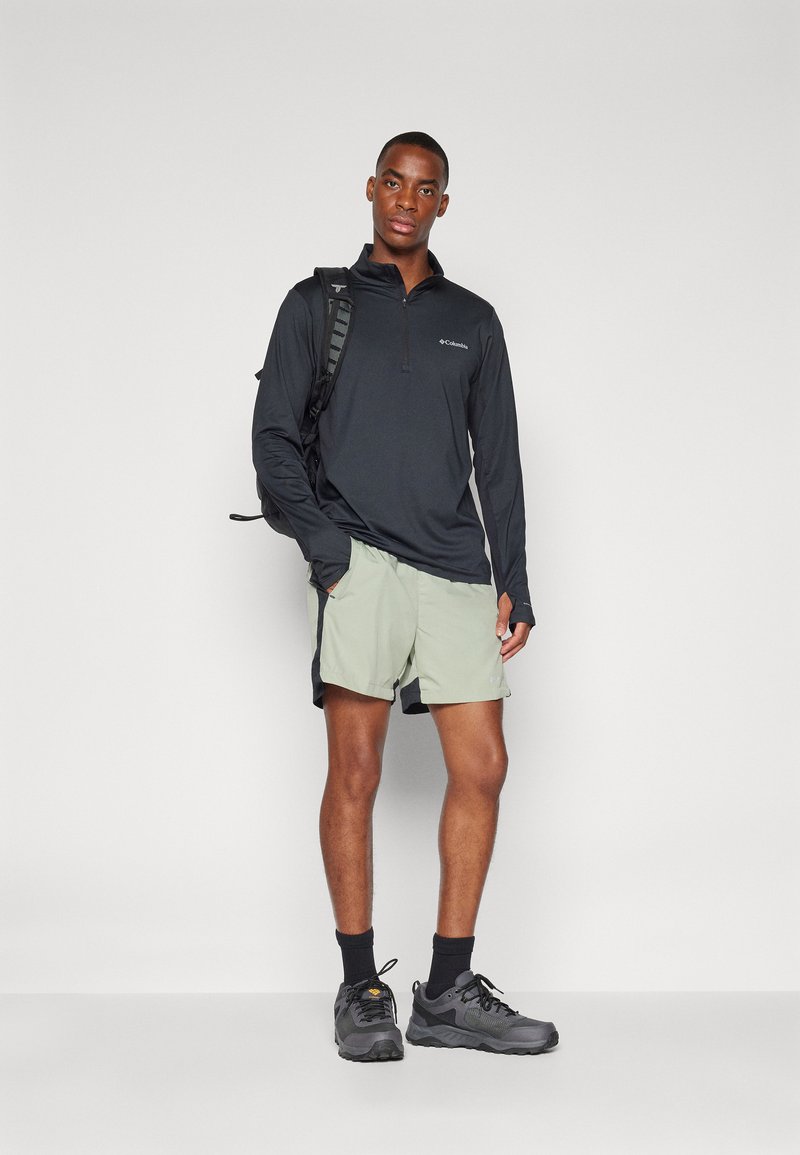 Top negro de manga larga con cremallera, pantalones cortos verdes claros, zapatos de senderismo grises y una mochila negra, situados frente a un fondo blanco liso.