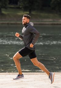 Athlet der løber forbi en vandmasse, iført en grå letvægtet hættejakke og sorte shorts, med grå og orange løbesko.