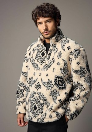 Fleece-Pullover in Creme mit schwarzen, aufwendigen Mustern, ausgestattet mit einem hohen Kragen und einem halben Reißverschluss. Weicher Stoff mit lockerer Passform.