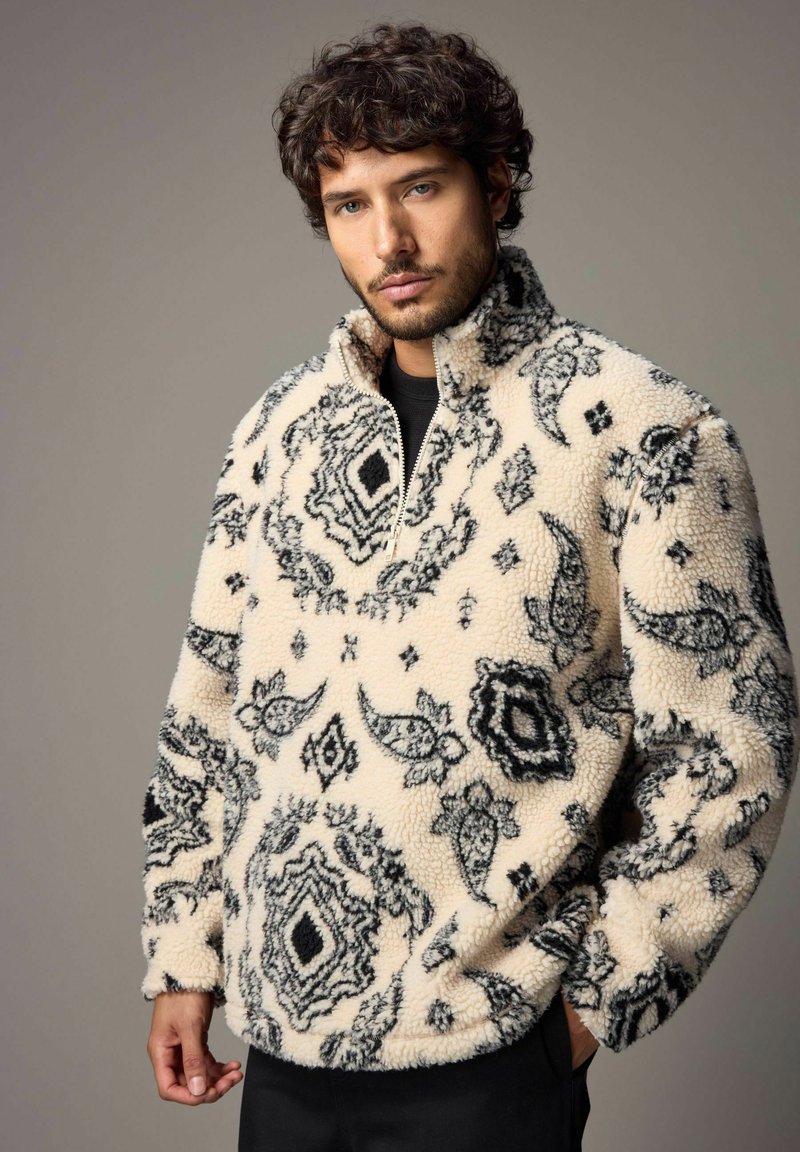 Fleece pullover i creme med sorte paisley-mønstre, halv-zip krave og struktureret overflade. Løs pasform og lange ærmer.