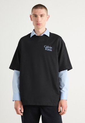 Black cotton t-shirt with a relaxed fit, featuring a light blue embroidered "Calvin Klein" logo on the chest, layered over a blue collared shirt.