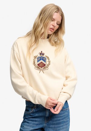 Lyshåret kvinde iført cremefarvet sweatshirt med broderet våbenskjold og blå jeans, kigger ned, mens hun justerer ærmet med begge hænder.