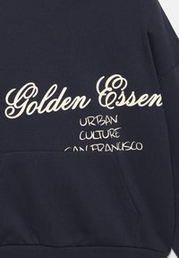 Sweat à capuche bleu marine avec poche avant, avec texte beige « Golden Essen » en écriture cursive et « Urban Culture San Francisco » en lettres majuscules.