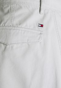 Gros plan sur un tissu blanc montrant une poche arrière avec un rabat et une petite broderie de logo rectangulaire rouge, blanc et marine.