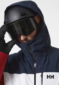 Veste de ski en blocs de couleurs bleu marine, rouge et blanc ; dotée d'un col haut, d'une fermeture éclair et d'un logo. Accompagnée d'un casque noir et d'une visière.