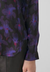 Mehrfarbige Bluse mit lila Blumenmustern auf schwarzem Hintergrund, mit einer glatten Textur und einem abgerundeten Kragen.