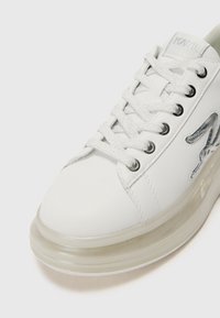 Zapatilla blanca de cuero con detalles plateados, puntera redonda, diseño de cordones y suela de goma transparente. Presenta un detalle de marca texturizado.