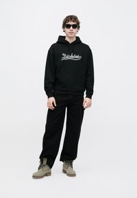 Μαύρο φούτερ με κουκούλα με το λογότυπο "Dickies", σε συνδυασμό με μαύρο παντελόνι και μπότες ελιάς. Το μοντέλο φοράει σκοτεινά γυαλιά ηλίου και στέκεται μπροστά σε λευκό φόντο.