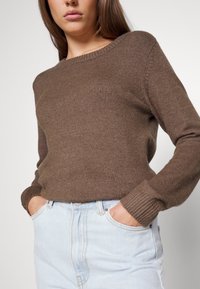Femme portant un pull en maille brun à manches longues et un jean taille haute bleu clair, les mains dans les poches, sur un fond uni.