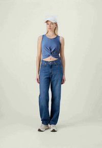 Top cropped azul com textura e detalhe torcido, combinado com jeans denim azuis largos. Usado com sapatilhas brancas e um boné branco. Fundo liso.
