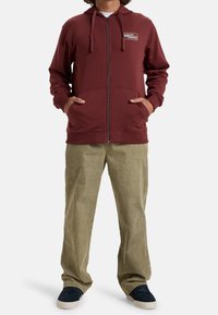 Sudadera con cremallera en color marrón con cordones, bolsillos frontales y logo de la marca. Combinada con pantalones de pana oliva claro y zapatos azul marino.