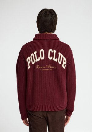 Persona que lleva un jersey de punto rojo oscuro con "POLO CLUB Beyond Classics Established 2012" bordado en la espalda, de espaldas.