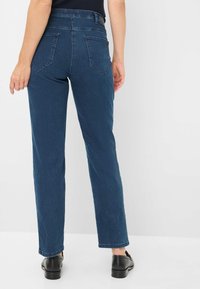 Jeans droits bleu marine avec taille haute, poches arrière et texture uni. Portés avec des chaussures noires sur un fond clair.