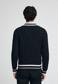 Svart kabelstickad tröja med vit randig krage, kontrasterande rand vid fållen, ribbade ärmar och en strukturerad mönstring.