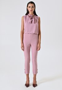 Blusa senza maniche rosa chiaro con scollo a fiocco, abbinata a pantaloni aderenti rosa chiaro con bottoni laterali e tacchi a punta marroni.