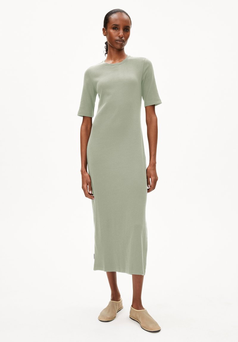 Geripptes, knielanges Kleid aus weichem grünem Stoff mit kurzen Ärmeln, ausgestattet mit einem runden Ausschnitt und seitlichen Schlitzen. Dazu werden beige Slipper getragen.