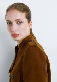 Veste en daim marron avec un col à revers, des poches latérales et des boutons décoratifs sur les épaules, sur un fond neutre.