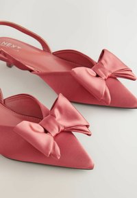 Escarpins en satin rose à dos ouverts, avec des pointes et de grands nœuds décoratifs à l'avant. Texture lisse et petit talon empilable.