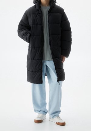 Persoon draagt een lange zwarte gewatteerde jas met capuchon, lichtblauwe wijde jeans, grijs T-shirt en witte sneakers met bruine zolen.