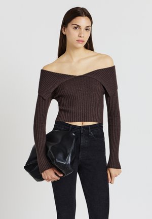 Braunes, geripptes, schulterfreies Crop-Top mit langen Ärmeln, kombiniert mit hoch taillierten schwarzen Jeans. Model hält eine große schwarze Ledertasche.