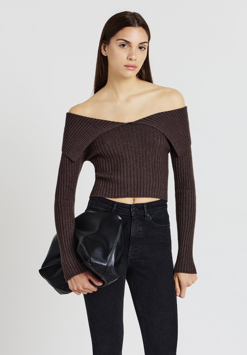 Braunes, geripptes, schulterfreies Crop-Top mit langen Ärmeln, kombiniert mit hoch taillierten schwarzen Jeans. Model hält eine große schwarze Ledertasche.