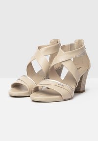Beige læder hælede sandaler med krydsede stropper, åben tå og en tekstureret blokhæle. Pyntet med sølvtrim på stropperne.