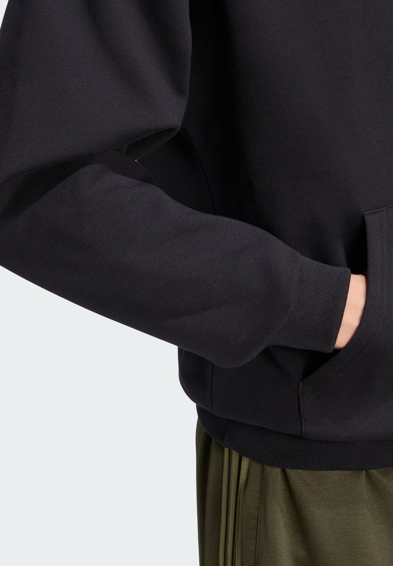 Personne portant un sweat-shirt noir à manches longues avec poignets côtelés et la main dans la poche avant, associé à un pantalon vert olive.