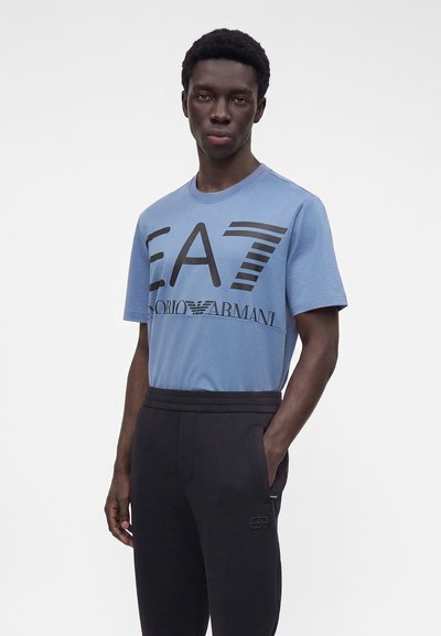 Млад мъж, носещ синя тениска EA7 Emporio Armani и черен панталон, стои с едната ръка в джоба пред обикновен фон.