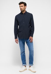 Camicia a maniche lunghe a quadri blu navy con bottone frontale, taschino al petto e polsini a contrasto, abbinata a jeans azzurri chiari e sneaker bianche.