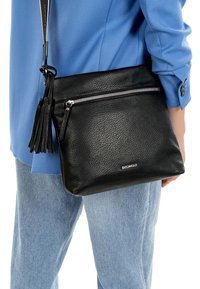 Person trägt hellblauen Blazer und Jeans und trägt eine schwarze, strukturierte Leder-Umhängetasche mit Reißverschluss und Quasten-Detail.