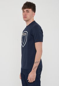 Mørkeblå bomulds t-shirt med et stort gråt skjoldlogo på forsiden. Korte ærmer og rund halsudskæring. Glat tekstur.