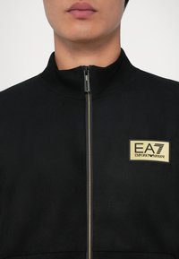 Primo piano di una persona che indossa una giacca nera con zip e una toppa beige del logo EA7 Emporio Armani sul petto.
