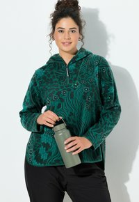 Zielona bluza polarowa na zamek z kwiatowymi i abstrakcyjnymi wzorami, długimi rękawami i zaokrąglonym dołem. Model trzyma matową zieloną butelkę na wodę.