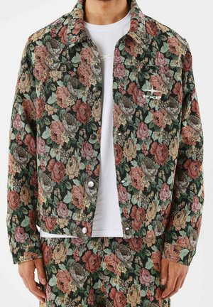 Man die een donker bloemenpatroon jasje en bijpassende broek draagt over een wit overhemd, staand met ontspannen handen langs het lichaam.