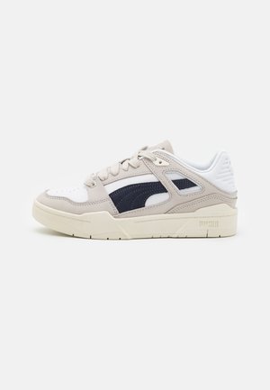 Beige ja valkoinen Puma-tennari mustalla raidallisella yksityiskohdalla, nauhallisella mallilla ja paksulla pohjalla valkoisella taustalla.