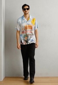 Kratke rokave, večbarvna srajca na gumb z grafiko znamenja motela in sonca, v kombinaciji z črnimi hlačami in temnimi sončnimi očali.