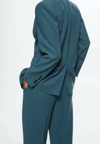 Modrozelený oblek s dobře střiženým sakem s rozparkem a černými knoflíkovými detaily, doplněný kalhotami se rovnými nohavicemi ze stejného materiálu a barvy.