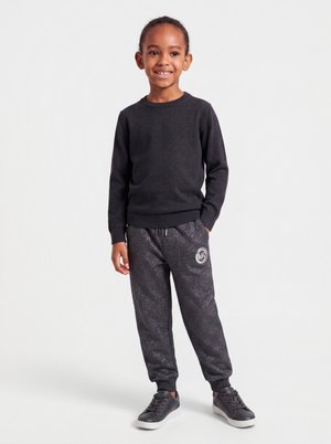 Jeune enfant debout et souriant, portant un tee-shirt noir à manches longues, un pantalon de jogging foncé tacheté avec logo, et des baskets noires à semelles blanches.