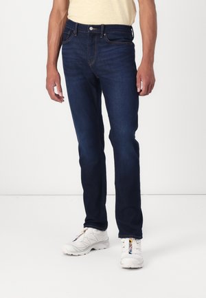 Jeans de mezclilla azul oscuro con un corte recto, que presentan detalles de costura sutiles y un diseño clásico de cinco bolsillos, combinados con zapatillas blancas.
