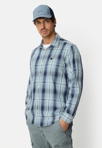 Hellblau kariertes Hemd mit Knopfleiste und Brusttasche, getragen mit einer blauen Kappe und kombiniert mit grauen Cargoshorts.