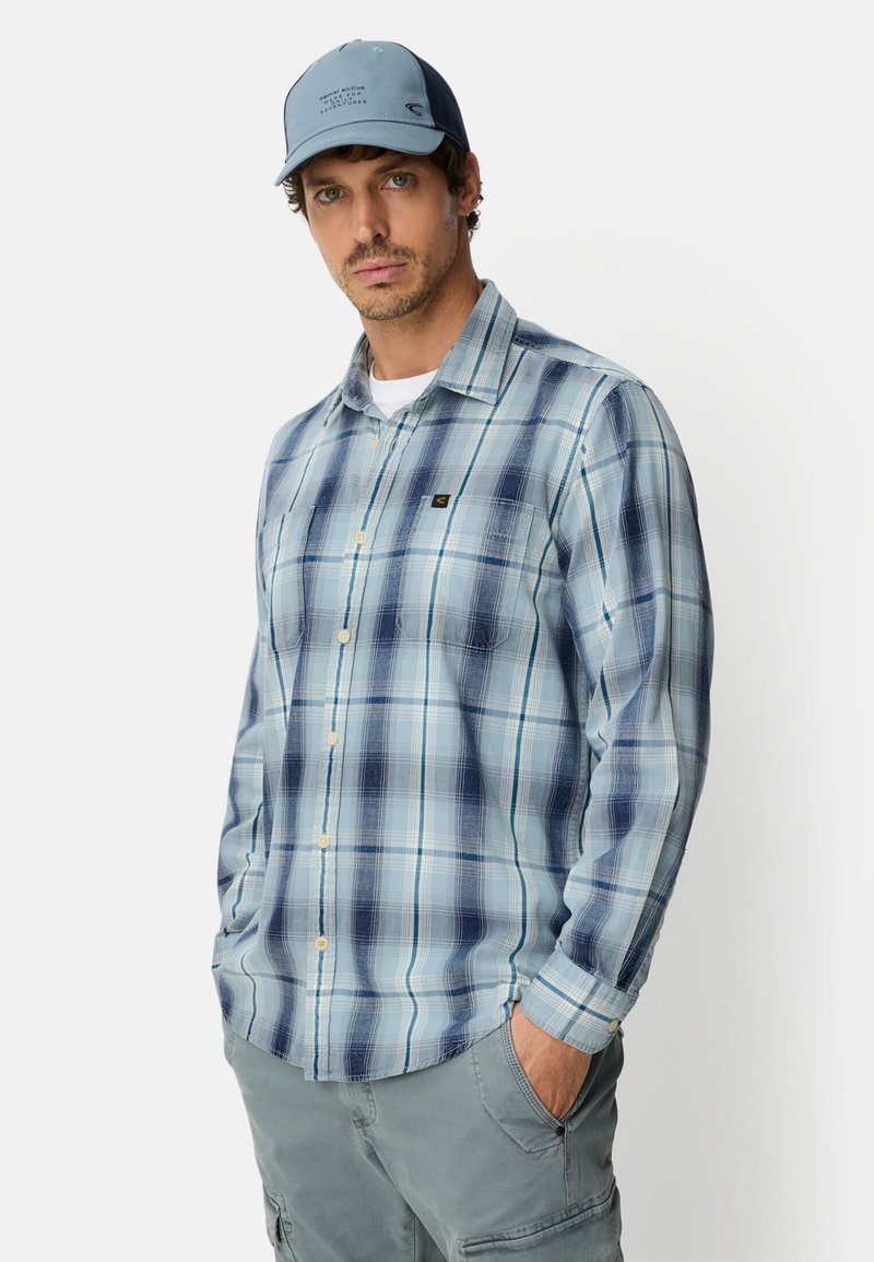 Hellblau kariertes Hemd mit Knopfleiste und Brusttasche, getragen mit einer blauen Kappe und kombiniert mit grauen Cargoshorts.