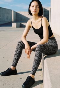 Tânără cu păr negru și scurt stă pe o margine de beton, purtând un maiou negru, colanți gri cu imprimeu leopard și adidași negri în aer liber.