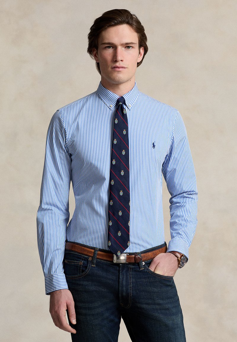 Modrá a biela pruhovaná košeľa s golierom na gombíky, doplnená tmavomodrou kravatou s malými emblémovými vzormi, nosená s tmavými džínsami a hnedým opaskom.