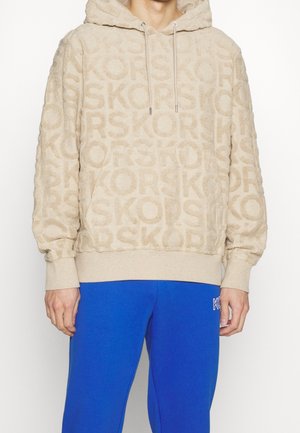 Beiger Kapuzenpullover mit geprägtem "KORS" Schriftzug, kombiniert mit leuchtend blauen Hosen mit einem kleinen "KORS" Logo am Oberschenkel.