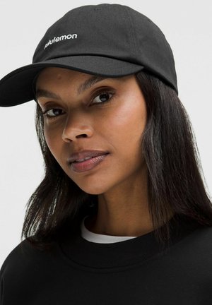 Mujer con cabello largo y oscuro, lleva una gorra negra de Lululemon y una camiseta negra, mirando ligeramente hacia un lado contra un fondo liso.