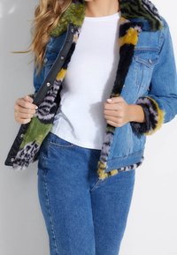 Chaqueta de mezclilla con un forro de piel sintética multicolor. Presenta patrones en verde, amarillo, negro y gris. Cierres de botón en el frontal y detalles en las mangas.
