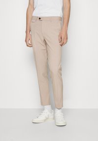 Pantaloni beige su misura, vestibilità slim, tessuto leggero, tasche frontali, chiusura con un solo bottone, indossati con sneakers bianche e calzini alla caviglia.